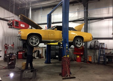 car repair in progress at Impact Fleet Service; it is a yellow car on a lift in the service garage with a mechanic in the background working on it